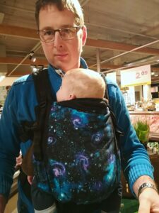 Op de foto zie je een papa met een kindje van 6 maanden in de Isara trendsetter draagzak met het galaxy motief. Papa draagt de baby op zijn buik met het gezicht van het kindje naar zich toe.